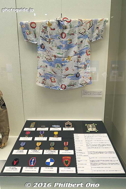 Aloha shirt for 100th Infantry Battalion veterans with a Japanese and Hawaiian design. The design includes torii, Japanese castle, shamisen, pagoda, pineapple, Gion Matsuri float, and "One Puka Puka" (100).
Below are military medals, badges, and patches.
