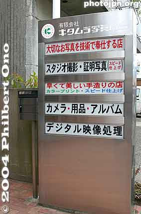 Photo Services
Sign outside a portrait studio and camera shop. 
From top to bottom: 
1st line from top (red characters): Irrelevant (ignore it) 
2nd line: Studio satsuei (portraits taken in the studio) and Shōmei shashin (ID photos). 
3rd line: Irrelevant 
4th line: Camera, yōhin (supplies), and album 
5th line: Digital eizō shori (Digital-editing services)
