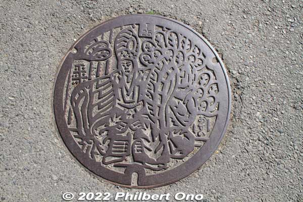 Hachioji manhole cover. It depicts a Sanbaso dancer (三番叟). Sanbaso is a well-known dancer in Noh and kabuki. He comes from a Noh prayer dance called Okina (翁) dating from the 14th century as a religious ritual. 
Hachioji's manhole shows the Sanbaso as a Hachioji Kuruma Ningyo puppet (八王子車人形). It's a unique offshoot of bunraku puppetry performed by a troupe based in Hachioji.
Here's a video that shows exactly what it is: https://youtu.be/XV8gVw5Rhwg
Keywords: tokyo hachioji takao manhole