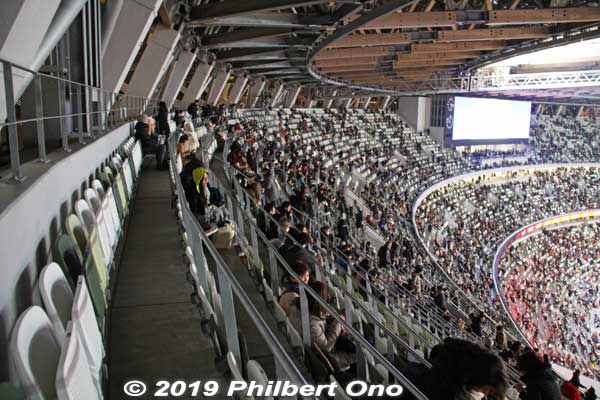 From the top row of seats in the stadium on the 3rd tier. Tier 3 has a steep 34˚ incline.
The Tier 1 is more gradual at 20˚, and Tier 2 is 29˚. Lateral space was minimized, so it's quite cramped to move through the seats when people are sitting in them. And there's no walkway traversing the seating area on the cramped 3rd tier. Go back out to the concourse to move to another block.
The stadium was designed this way to keep the seats closer to the field. I must say, it is effective in that sense. The field looks closer and you get clear views with no heads in the way. In the old National Stadium, the top row was faraway from the field.
This top row is also right below the open-air part of the stadium and if it's windy and rainy, I would think people may get wet here. Must be cold in winter to sit here. But in summer, it might be cooler when there's a breeze.
Keywords: tokyo shinjuku olympic national stadium